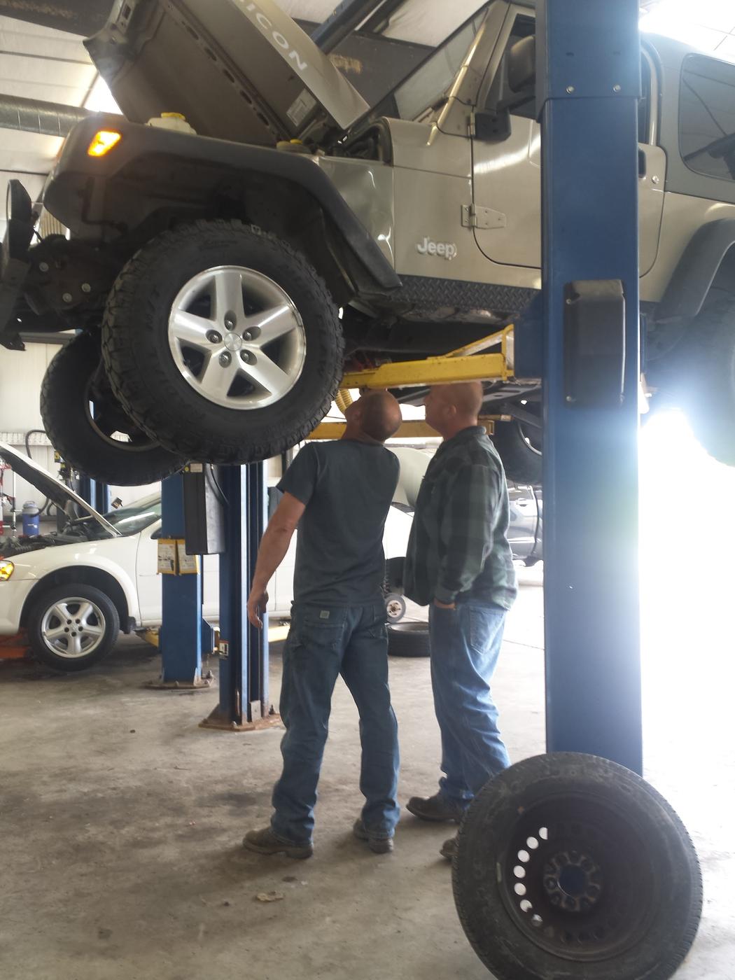A technician and a customer stood beneath a Jeep raised on a lift, both looking up at the underside